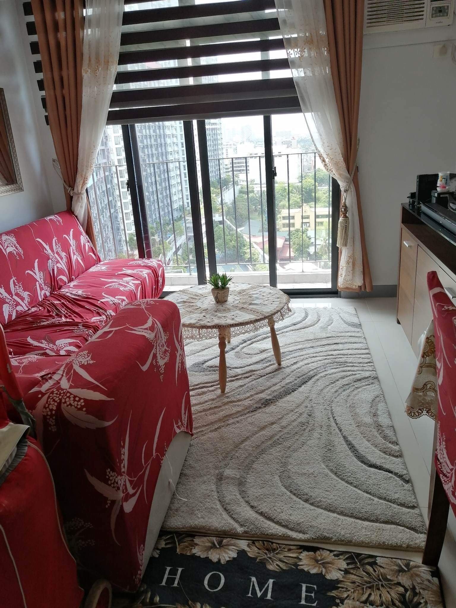 Sala de estar com um sofá vermelho, uma mesinha redonda com uma planta, e vista para edifícios através de grandes janelas.