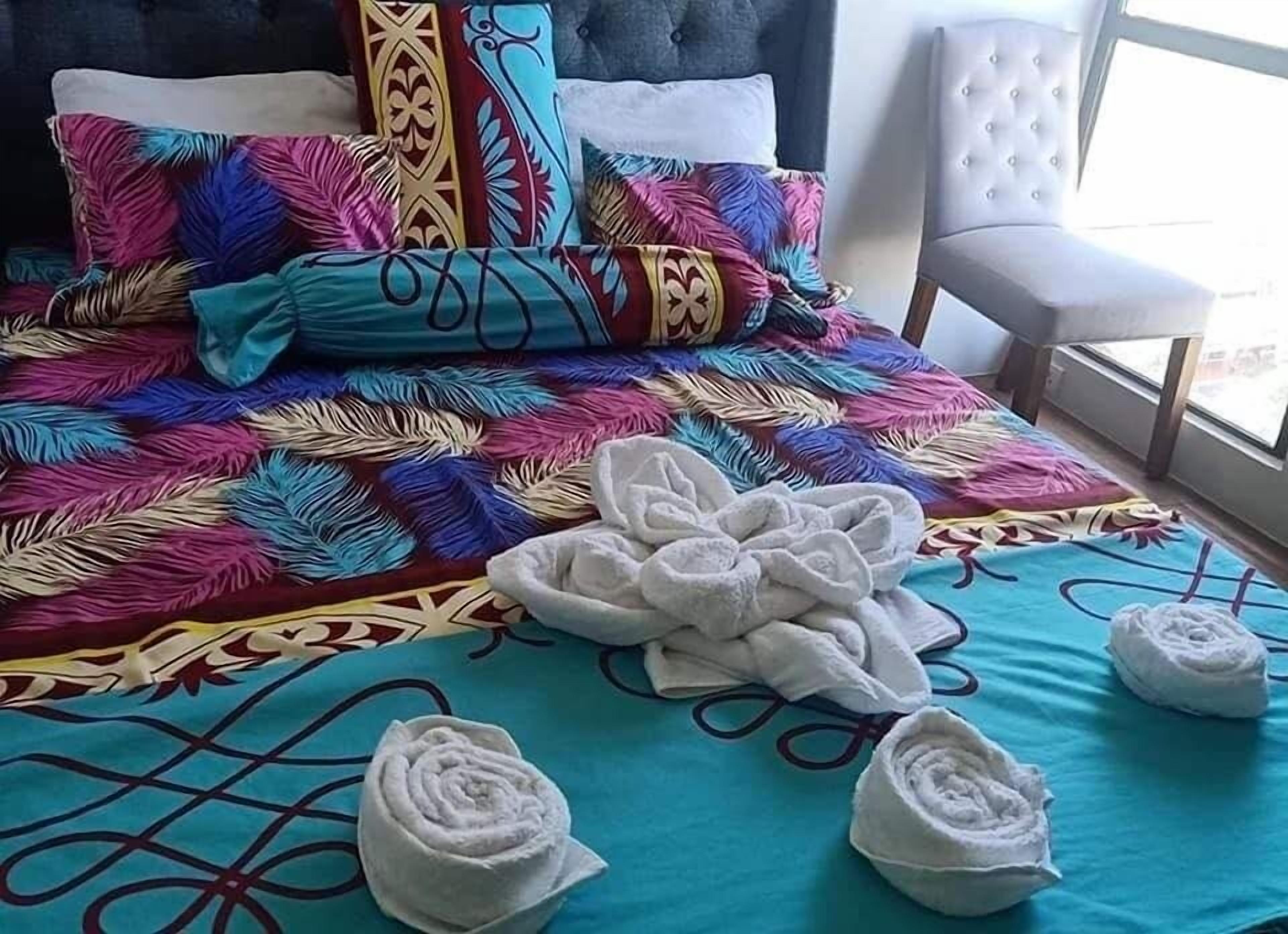 Een bed met kleurrijke kussens en een blauwe deken, een witte stoel en opgerolde handdoeken op het bed.