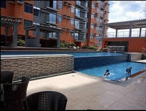 A swimming pool area with a stone wall, outdoor seating, and a multi-story building in the background.