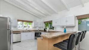 A modern kitchen with white cabinets, a granite countertop, a stainless steel refrigerator, a dishwasher, and a bar area with black bar stools. - 3BR 2.5BA Villa w/Pool in Palm Beach (Noord)
