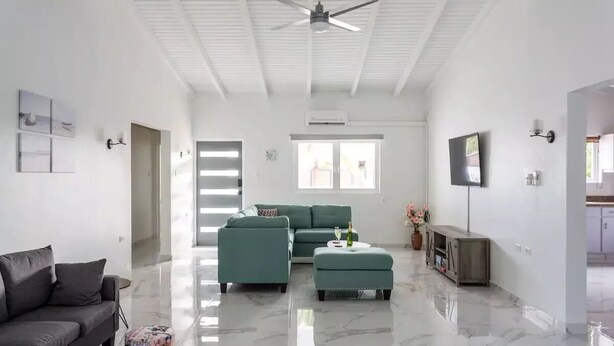 A spacious living room with a green sectional sofa, a grey couch, and a small ottoman. There is a ceiling fan, a flat-screen TV, and a marble floor. - 3BR 2.5BA Villa w/Pool in Palm Beach (Noord)