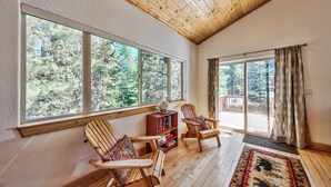 A spacious room with large windows, wooden flooring, and a cozy seating area with two chairs and a bookshelf. - Alpine Family Adventure - Pets, BBQ, Games (South Lake Tahoe)