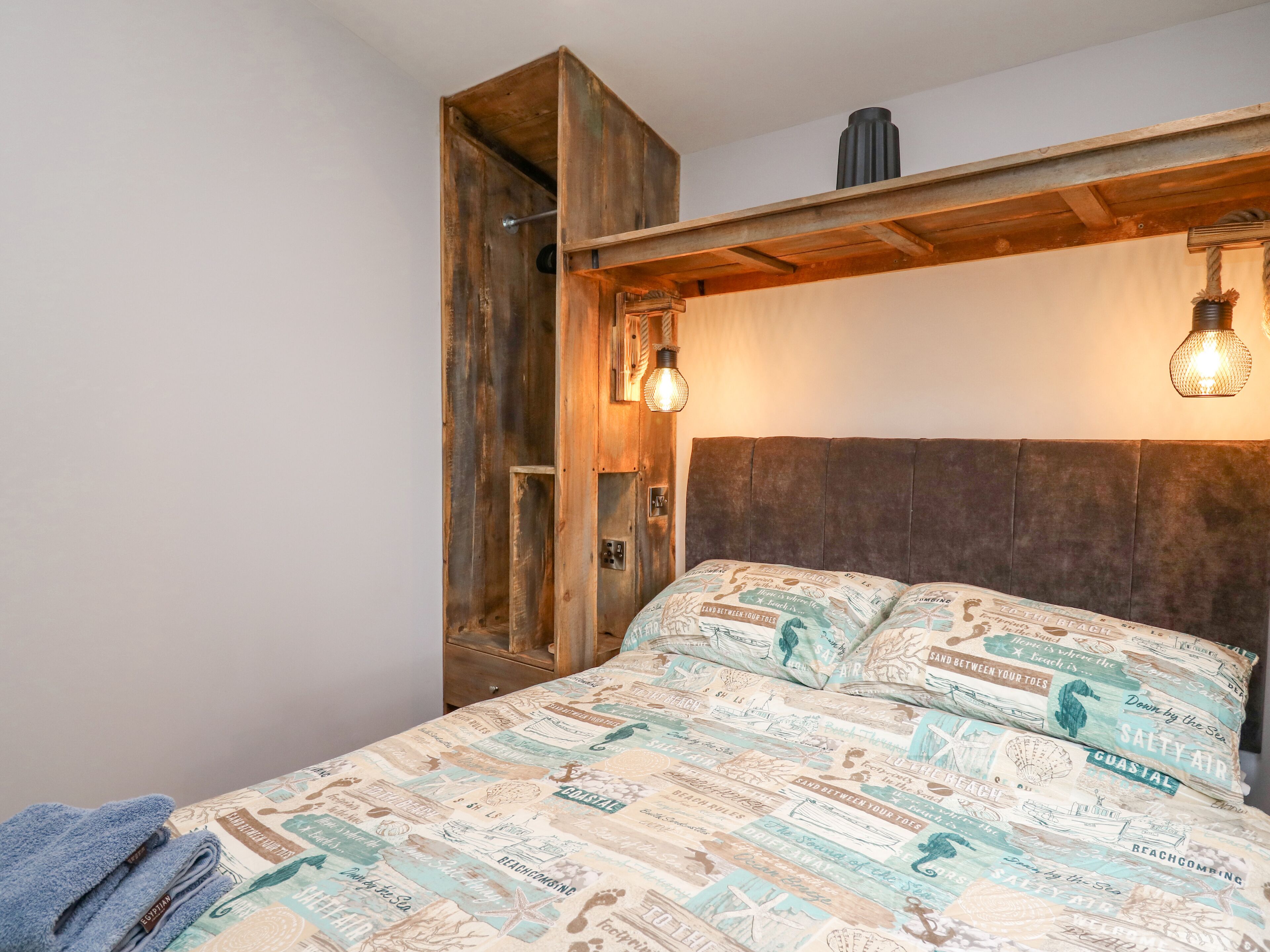 A bed with a coastal-themed duvet, a wooden headboard, and a hanging light fixture.