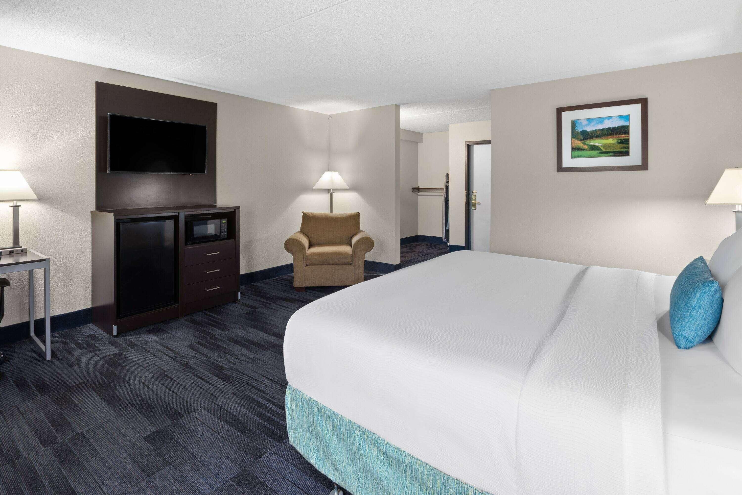 A hotel room with a bed, a TV mounted on the wall, a desk with a lamp, a chair, and a door leading to another room.