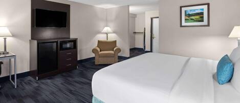 A hotel room with a bed, a TV mounted on the wall, a desk with a lamp, a chair, and a door leading to another room.