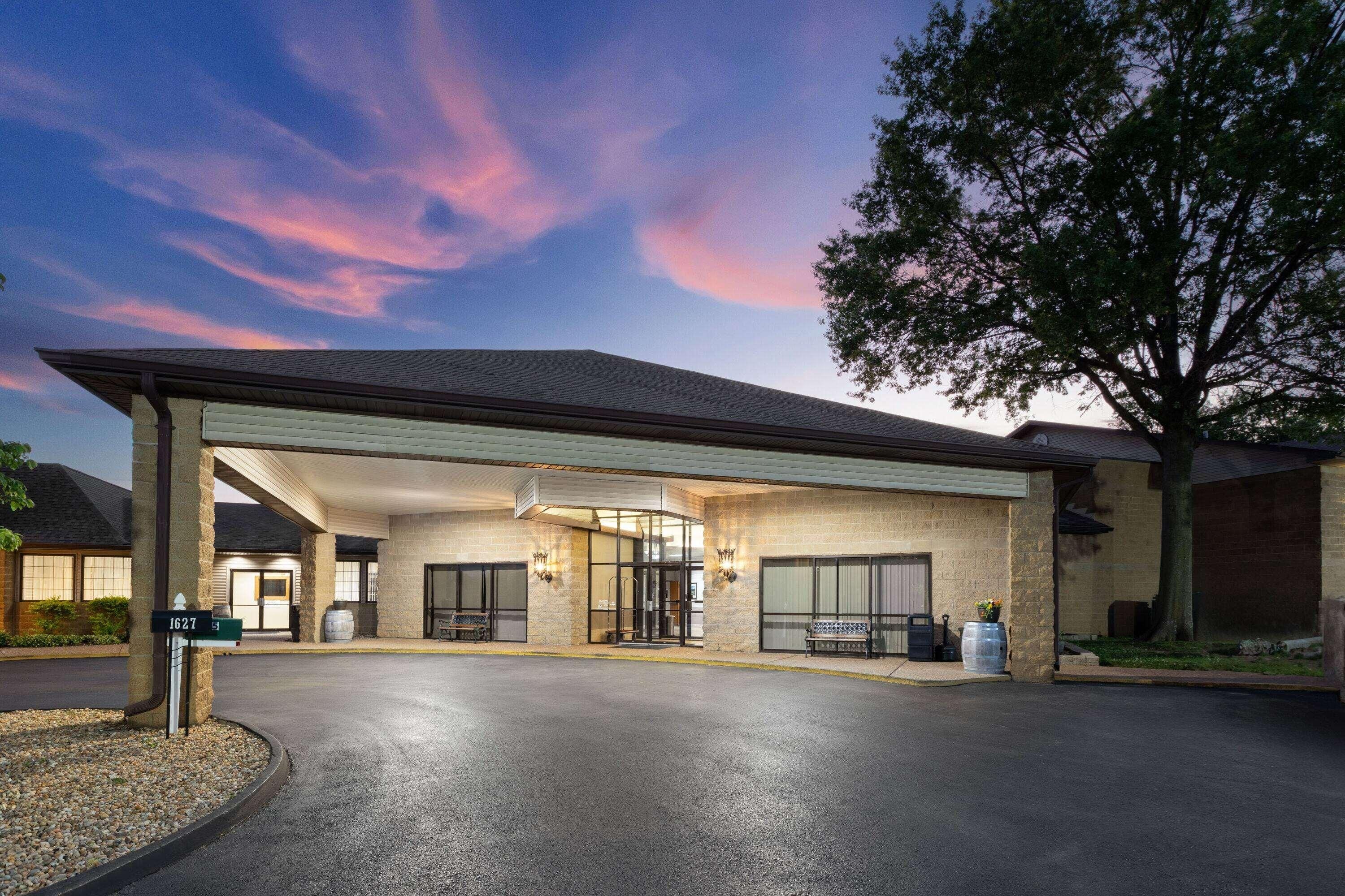 A hotel entrance with a covered driveway, a sign indicating the address, and a large tree to the right.