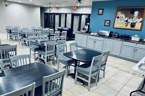 A dining area with multiple tables and chairs, a kitchen area with cabinets and appliances, and a painting on the wall. - Baymont Inn And Suites By Wyndham Farmington Mo (Farmington)