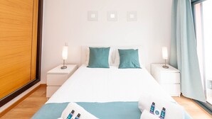A neatly arranged hotel room with a large bed, two bedside tables with lamps, a wooden wardrobe, and a view of the city through the window. - Almond Apartment Ocv - Pool & Garage (Albufeira)