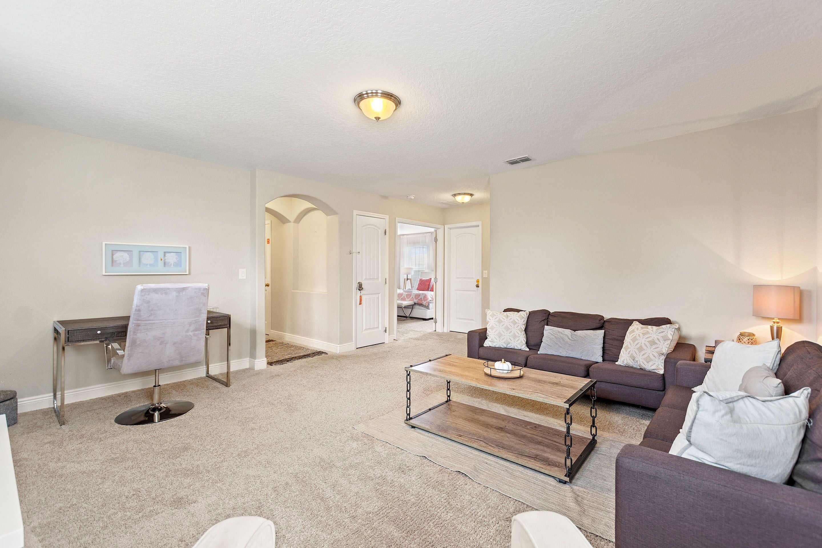 A living room with a sofa, coffee table, and a chair. There is a desk with a chair and a lamp. The room has a carpeted floor and white walls.
