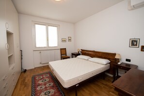 Un dormitorio con una cama de madera, una mesita de noche, un escritorio, un ventanal y un radiador.