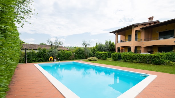 A rectangular swimming pool with a tiled deck, surrounded by well-maintained greenery and a two-story building with balconies. - Casa della Rocca (Manerba del Garda)