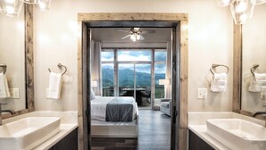A hotel room with a large bed, two sinks, a ceiling fan, and a view of mountains through large windows.