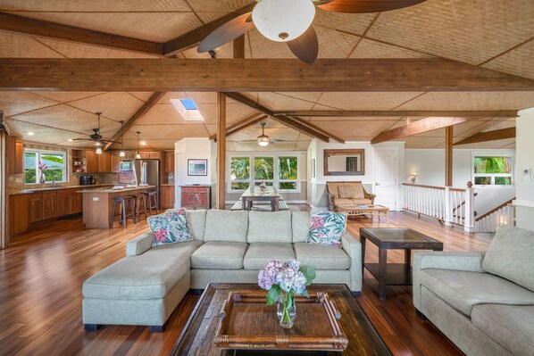 A spacious living area with a large sectional sofa, a coffee table with a vase, wooden flooring, and a kitchen in the background.