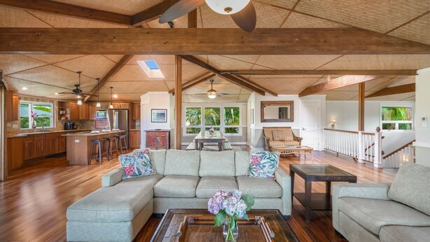 A spacious living area with a large sectional sofa, a coffee table with a vase, wooden flooring, and a kitchen in the background. - Lola Hale, Spacious Upgraded Hawaiiana House, A/C and Hot Tub (Princeville)