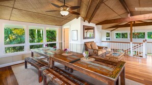 A spacious room with a wooden dining table, benches, and a ceiling fan. Large windows provide views of greenery outside. - Lola Hale, Spacious Upgraded Hawaiiana House, A/C and Hot Tub (Princeville)