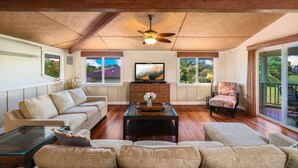 A spacious living room with a high ceiling, wooden beams, a ceiling fan, a large flat-screen TV, a sofa, a coffee table, and a chair. - Lola Hale, Spacious Upgraded Hawaiiana House, A/C and Hot Tub (Princeville)