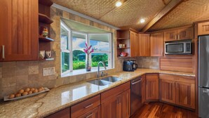 A kitchen with wooden cabinets, a granite countertop, a stainless steel refrigerator, and a window with a view of greenery. - Lola Hale, Spacious Upgraded Hawaiiana House, A/C and Hot Tub (Princeville)