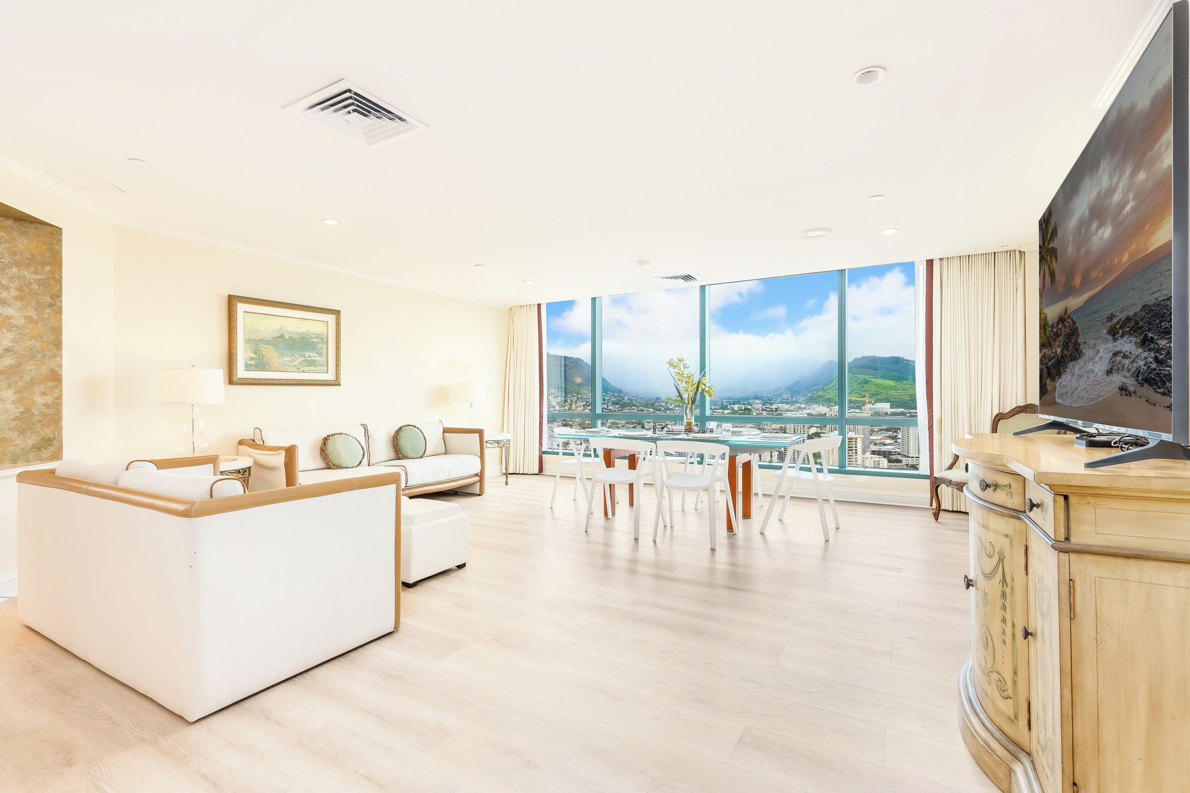 A modern hotel room with a large window, a sofa, a dining table, and a TV.
