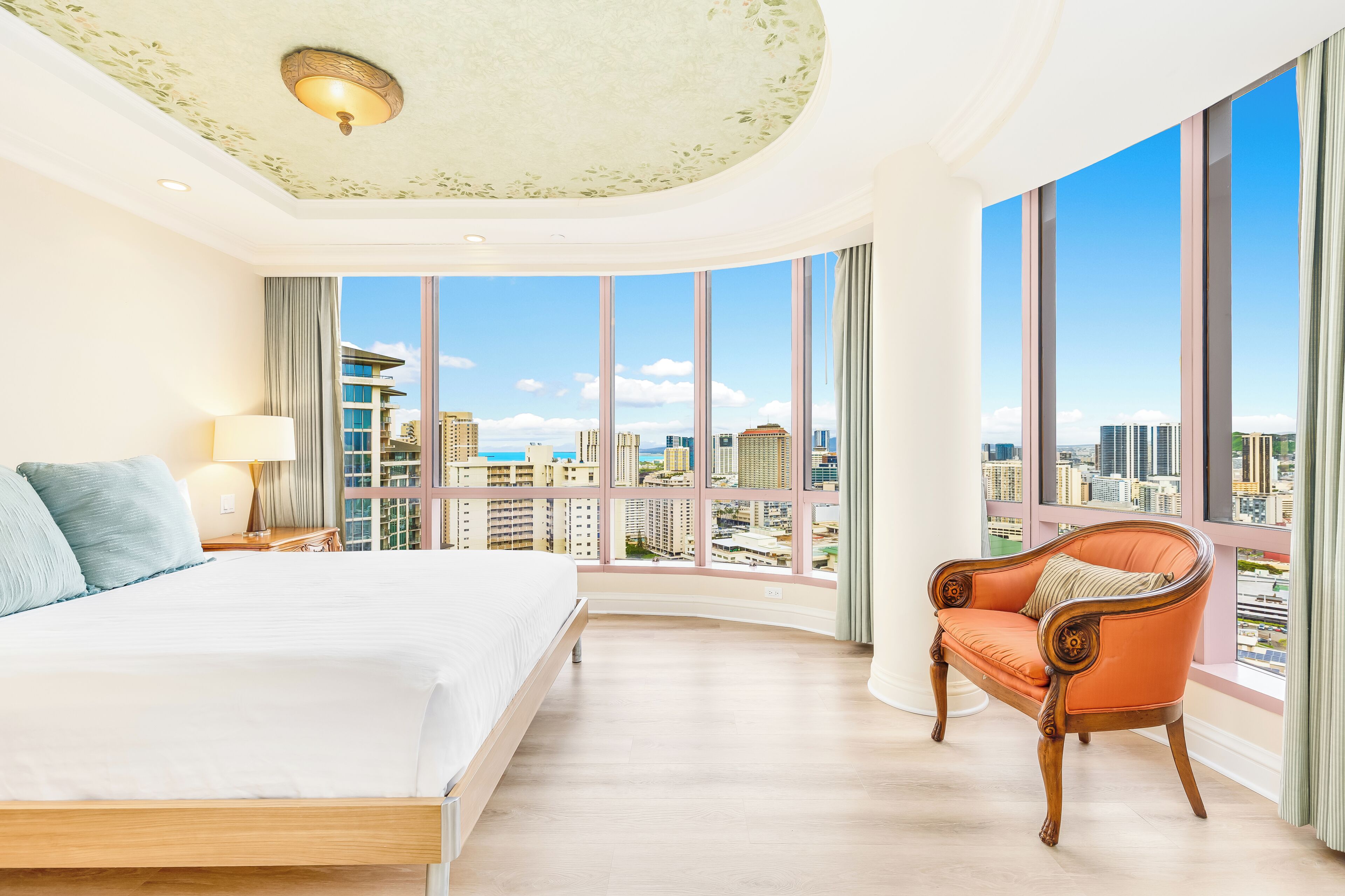 A hotel room with a large bed, a chair, a nightstand, and a view of the cityscape through large windows.
