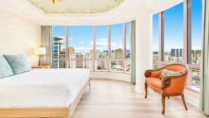 A hotel room with a large bed, a chair, a nightstand, and a view of the cityscape through large windows.