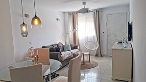 Ein modernes Wohnzimmer mit einem Glastisch, weißen Stühlen, einem grauen Sofa und einem weißen Sessel. Es gibt einen Deckenventilator, eine weiße Kommode und ein an der Wand montiertes Fernsehgerät.