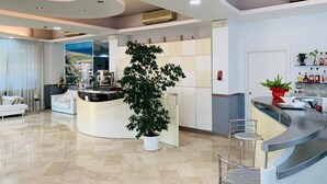 A modern hotel lobby with a curved reception desk, a large potted plant, and a seating area with a white sofa and chairs. - Hotel Marittima (Rimini)