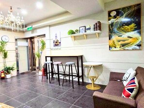 Un hall de hotel con mostrador de recepción, taburetes de bar, sofá, candelabro, reloj y un cuadro en la pared.