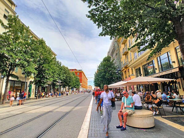 Eine belebte Straßenszene mit Straßenbahnschienen, Fußgängern und Außenbereichsgastronomie.