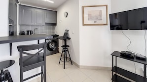 Une cuisine compacte avec des meubles gris, un lave-linge, un petit tabouret de bar et un tableau encadré au mur.