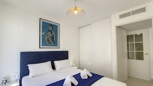 Un lit bien fait, avec une literie blanche et bleue, une œuvre d’art encadrée au-dessus et une suspension moderne.