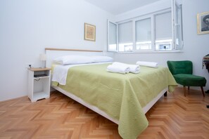 Ein ordentlich gemachtes Bett mit einer grünen Tagesdecke und weißen Kissen, ein Nachttisch mit einer Lampe, ein grüner Sessel und ein Fenster mit Jalousien.