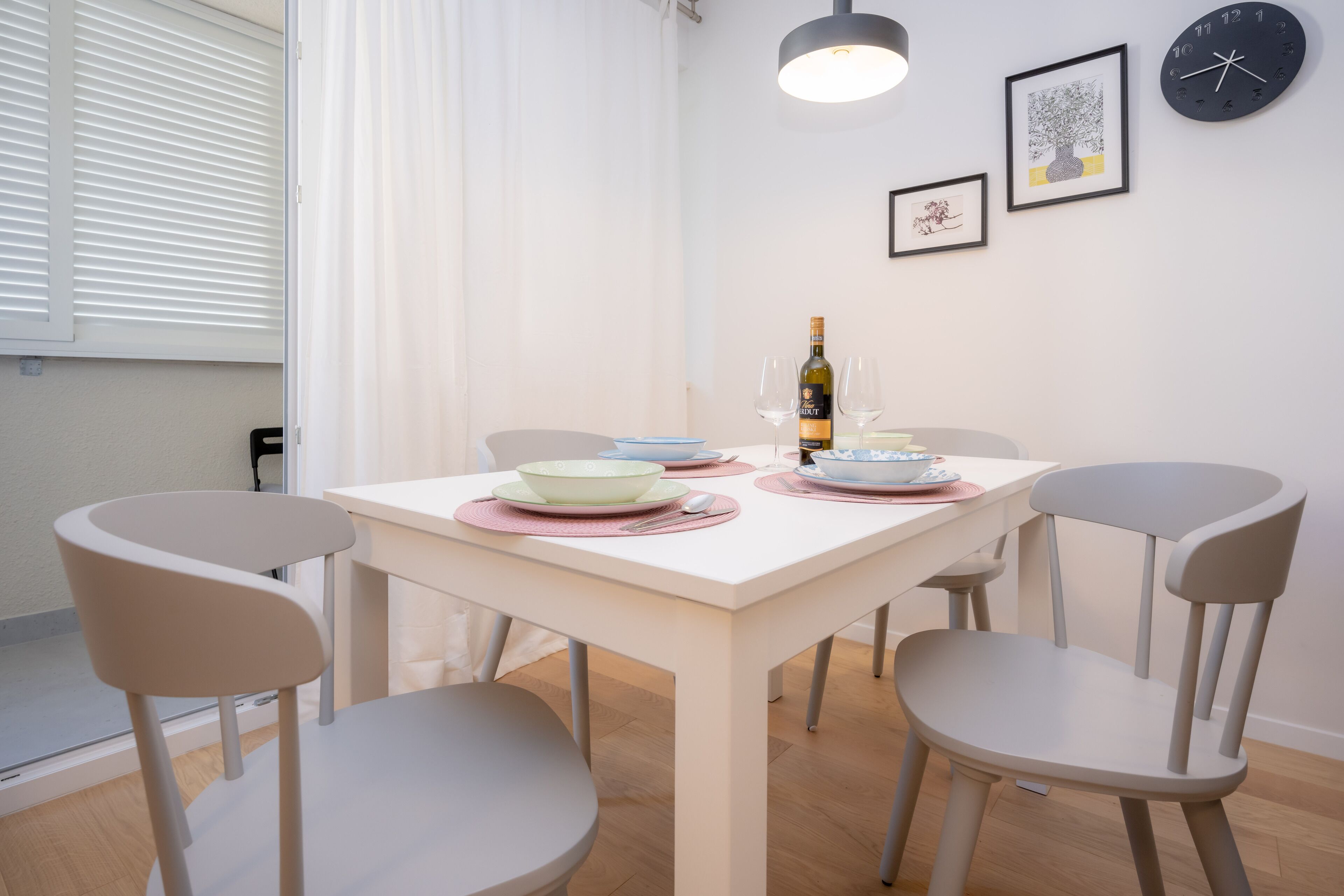 A dining area with a white table, four grey chairs, a bottle of wine, and two glasses. There are framed pictures on the wall and a clock.