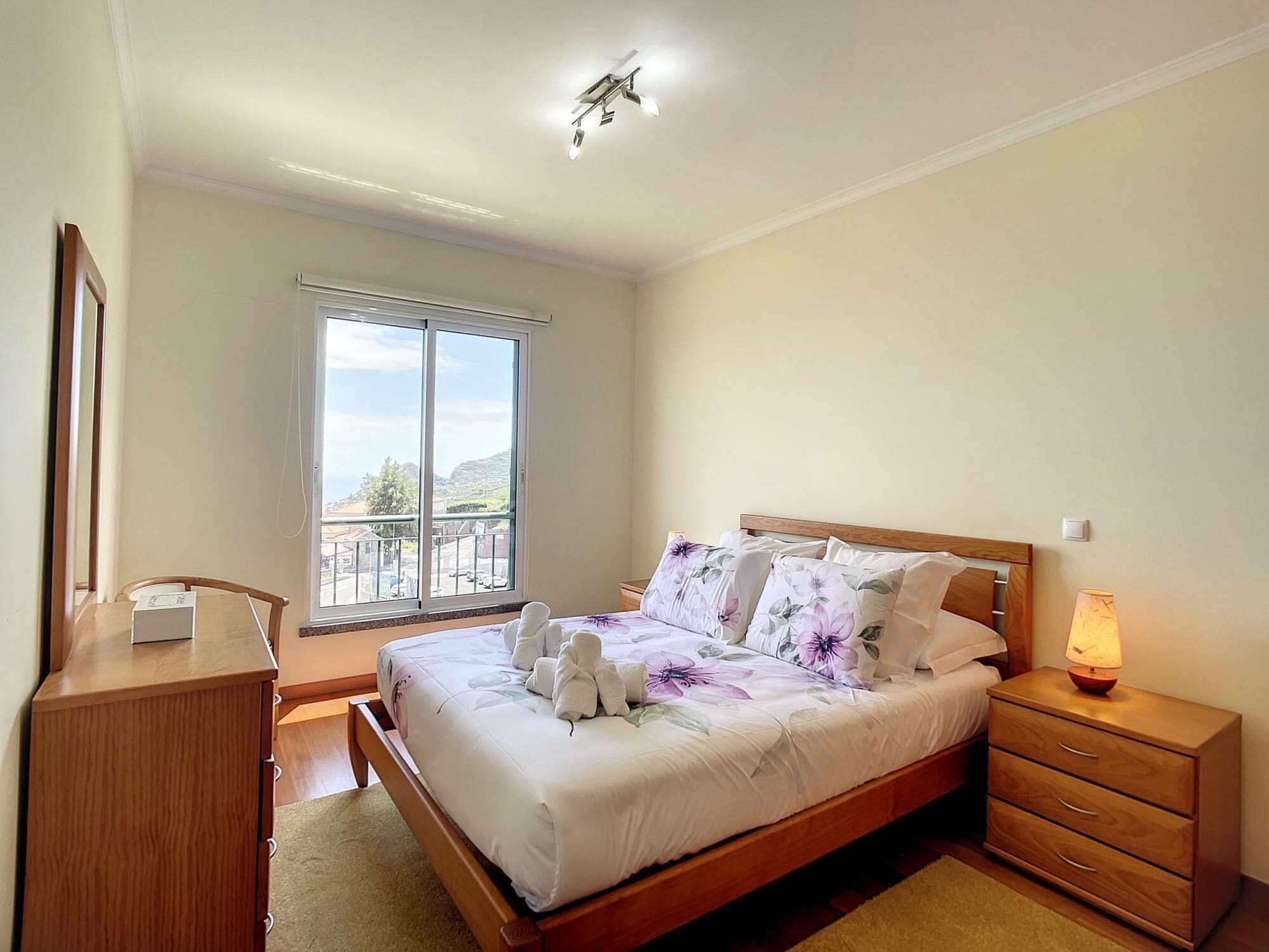 A bedroom with a bed, bedside table, lamp, and a view of the outside.