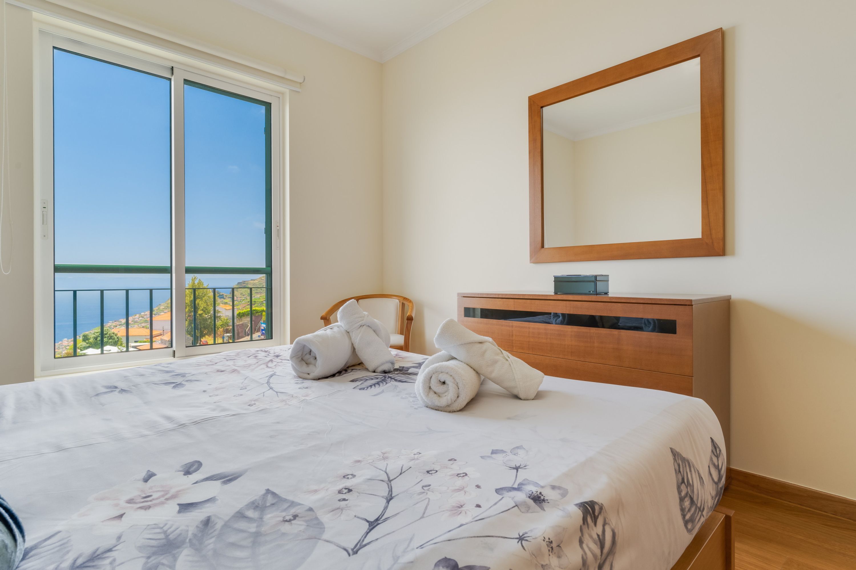 A bedroom with a bed, a wooden dresser, a mirror, and a view of the sea through a sliding glass door.