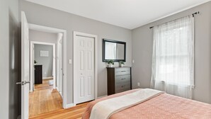 A bedroom with a wooden floor, a bed with a patterned bedspread, a dresser, a mirror, and a door leading to another room.