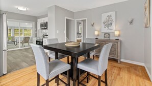 A dining area with a black table and grey chairs, a kitchen in the background, and a grey wall with a framed picture.