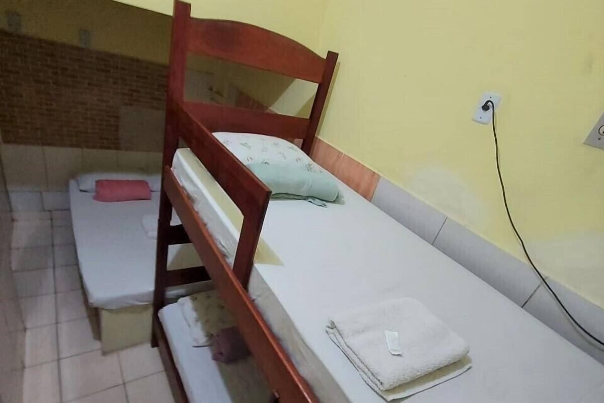 A single bed with a wooden frame and a white mattress, a small bedside table with a pink pillow, and a wall socket.