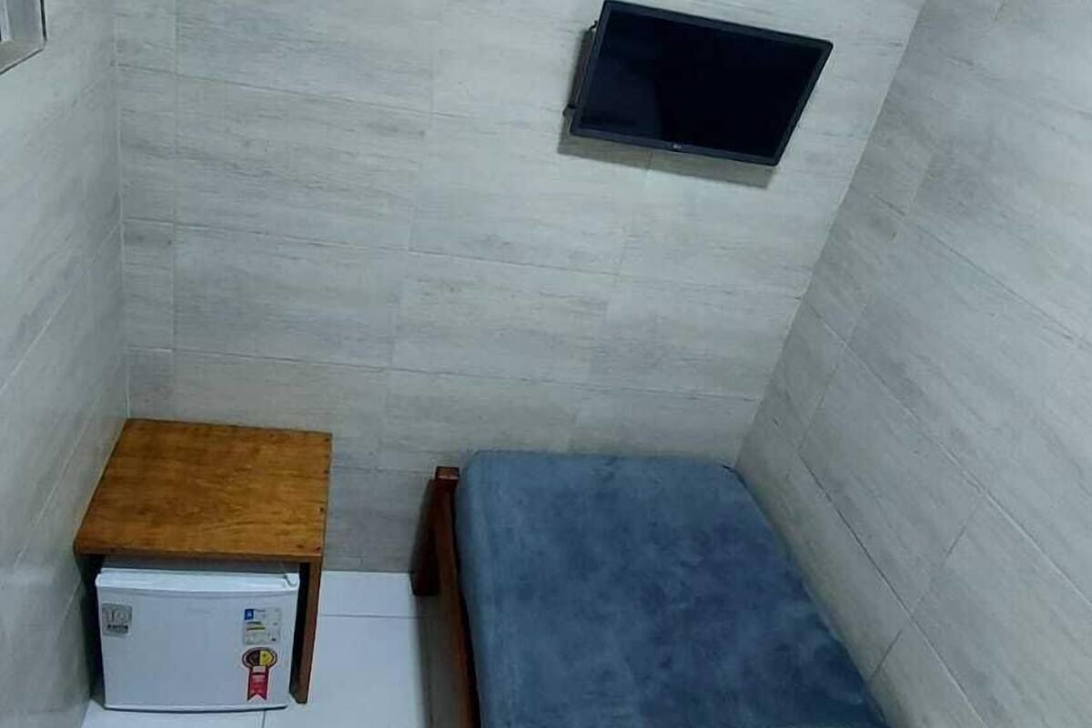 A small, compact room with a bed, a small table, a TV mounted on the wall, and a refrigerator.