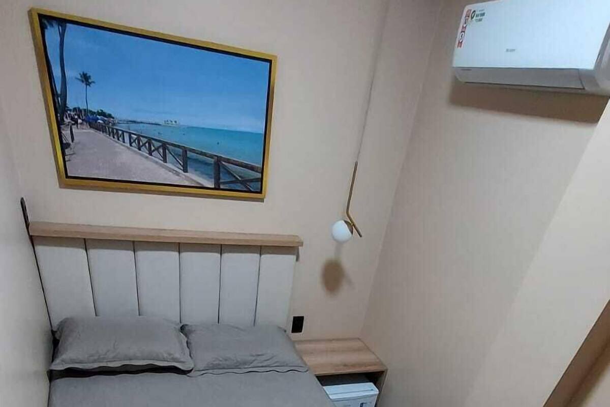 A neatly made bed with a beige blanket and a heart-shaped pillow, a painting of a beach scene on the wall, and an air conditioning unit mounted on the wall.