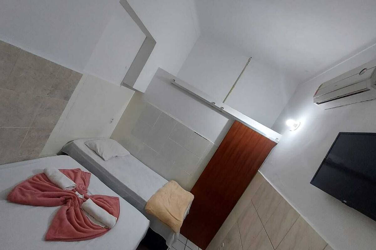A hotel room with a bed, a small table, a TV, and an air conditioning unit.