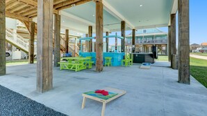 Ein überdachter Patio-Bereich im Freien mit Holzbalken, grünen Tischen und Stühlen sowie einem Spieltisch aus Holz mit einem roten Gegenstand darauf.