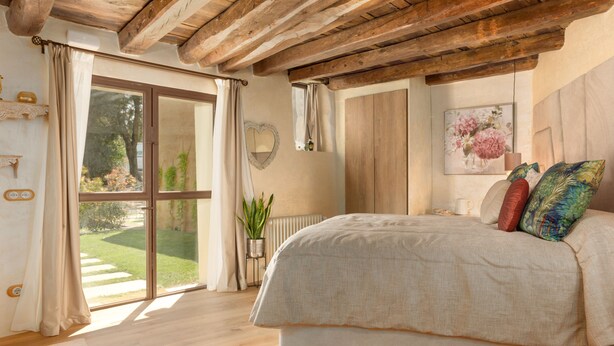 Une chambre avec un grand lit, des poutres apparentes en bois au plafond, une vue sur un jardin accessible par une porte coulissante et une plante en pot.