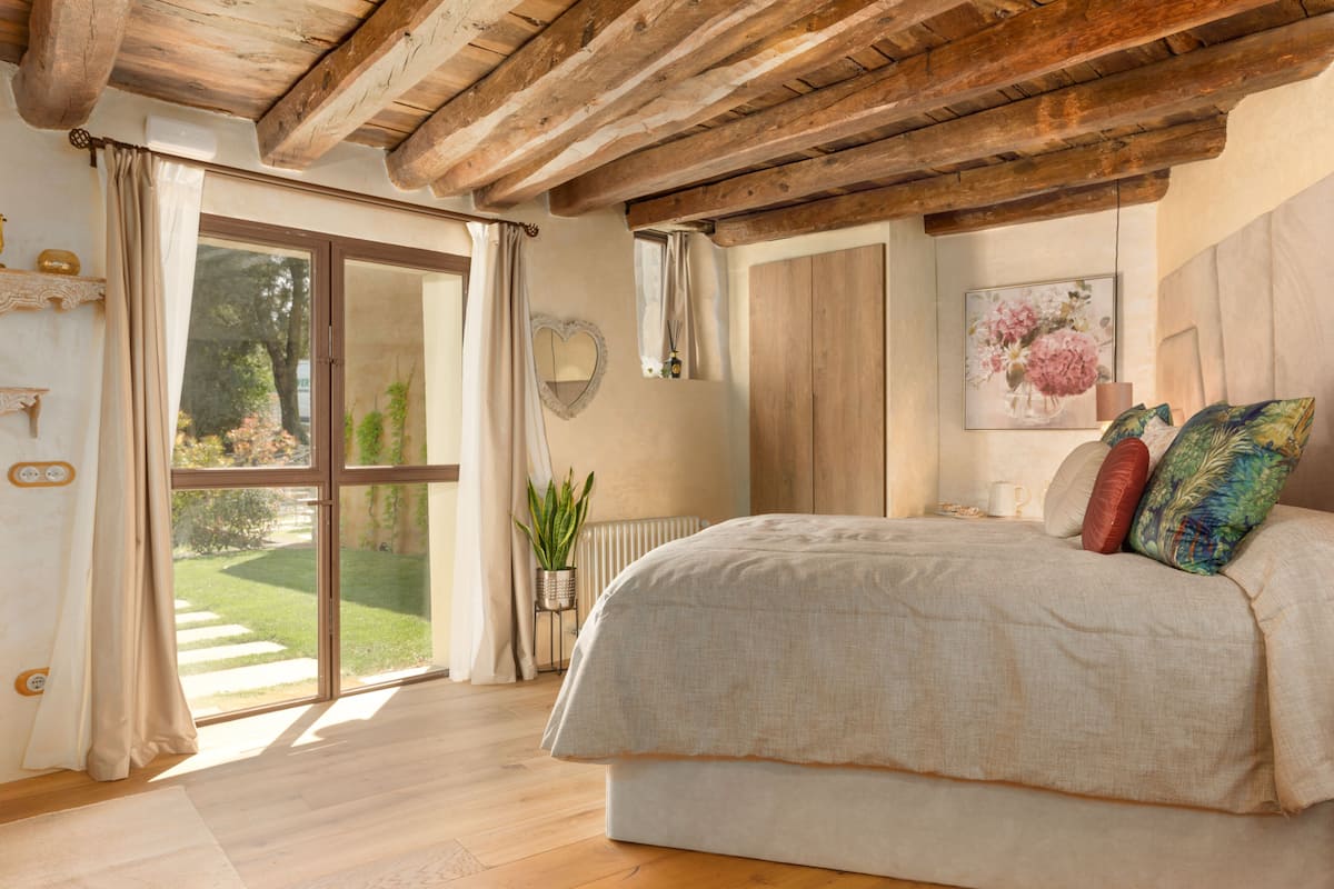 Un dormitorio con una cama grande, vigas de madera en el techo, vista a un jardín por una puerta corredera y una planta en maceta.