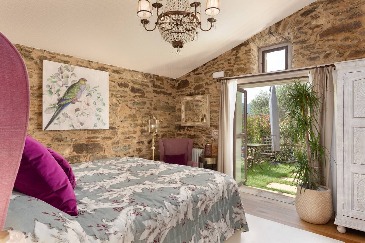 Un dormitorio con pared de piedra, una cama con cubrecama floral, un candelabro, un ventanal grande con cortinas y vista a un jardín.