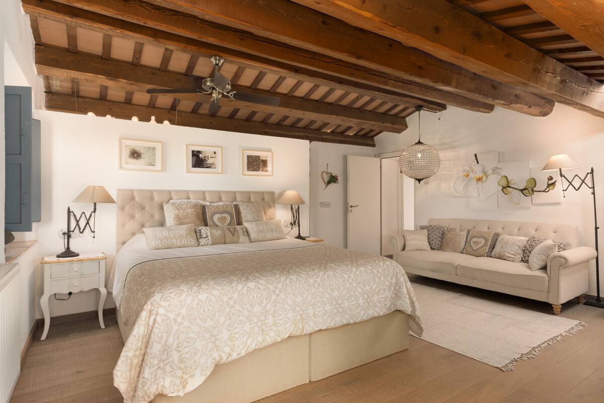 Un dormitorio con una cama grande, un sofá, un ventilador de techo y obras de arte colgadas en la pared.