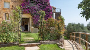 Un bâtiment en pierre avec une entrée donnant sur un jardin, un pont en bois et un escalier menant à l’entrée.