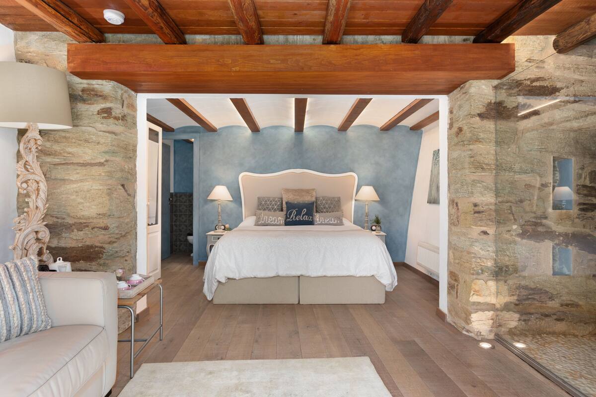Un dormitorio con una cama grande, paredes de piedra, vigas de madera y una mesita de noche con lámpara.