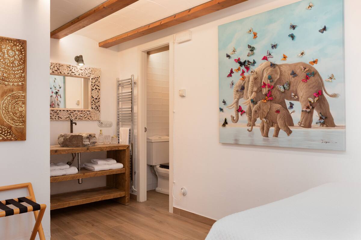 Un baño con tocador de madera, un espejo con detalles tallados, un perchero y un cuadro con elefantes y mariposas.