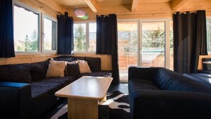 A cozy cabin interior with a wooden ceiling, a sofa, a table, and large windows offering a view of the outdoors.