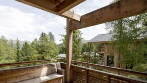 A wooden deck with a bench and railing, overlooking a forested area with a building in the background.